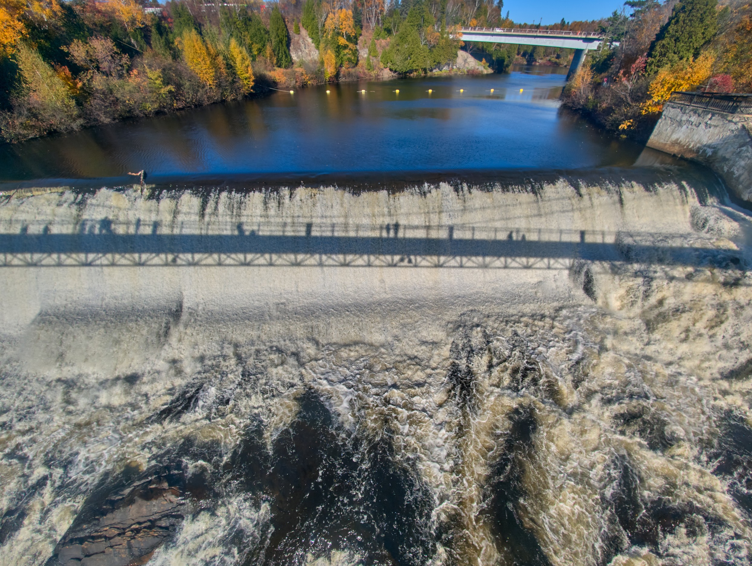 Vermont and Quebec 43 An elevated view shows a horizontal concrete barrier spanning dark river water between autumn-covered rocky banks.