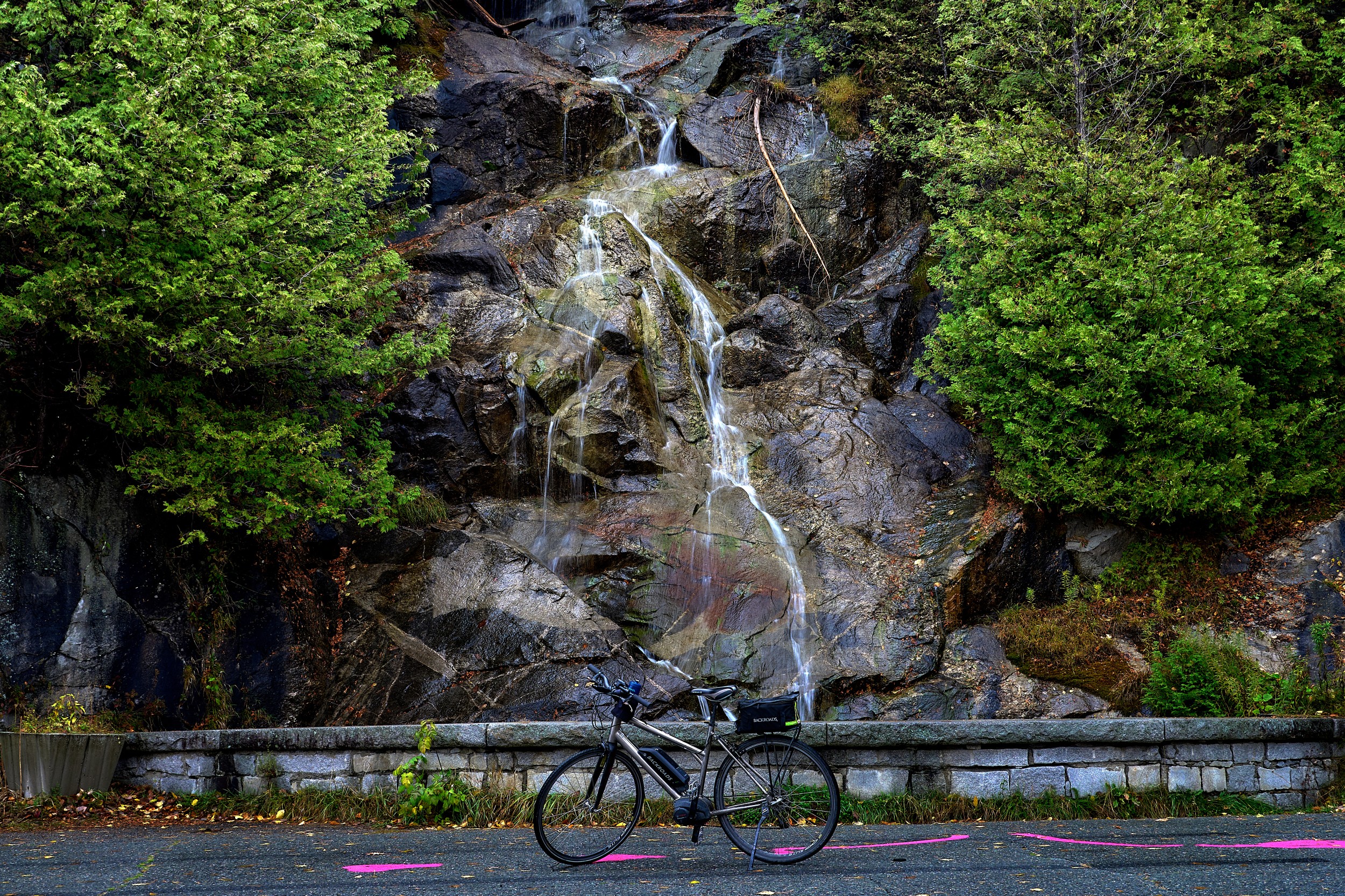 Vermont and Quebec 81 Narrow waterfall runs down dark rock behind a roadside guardrail with a black bicycle in front.