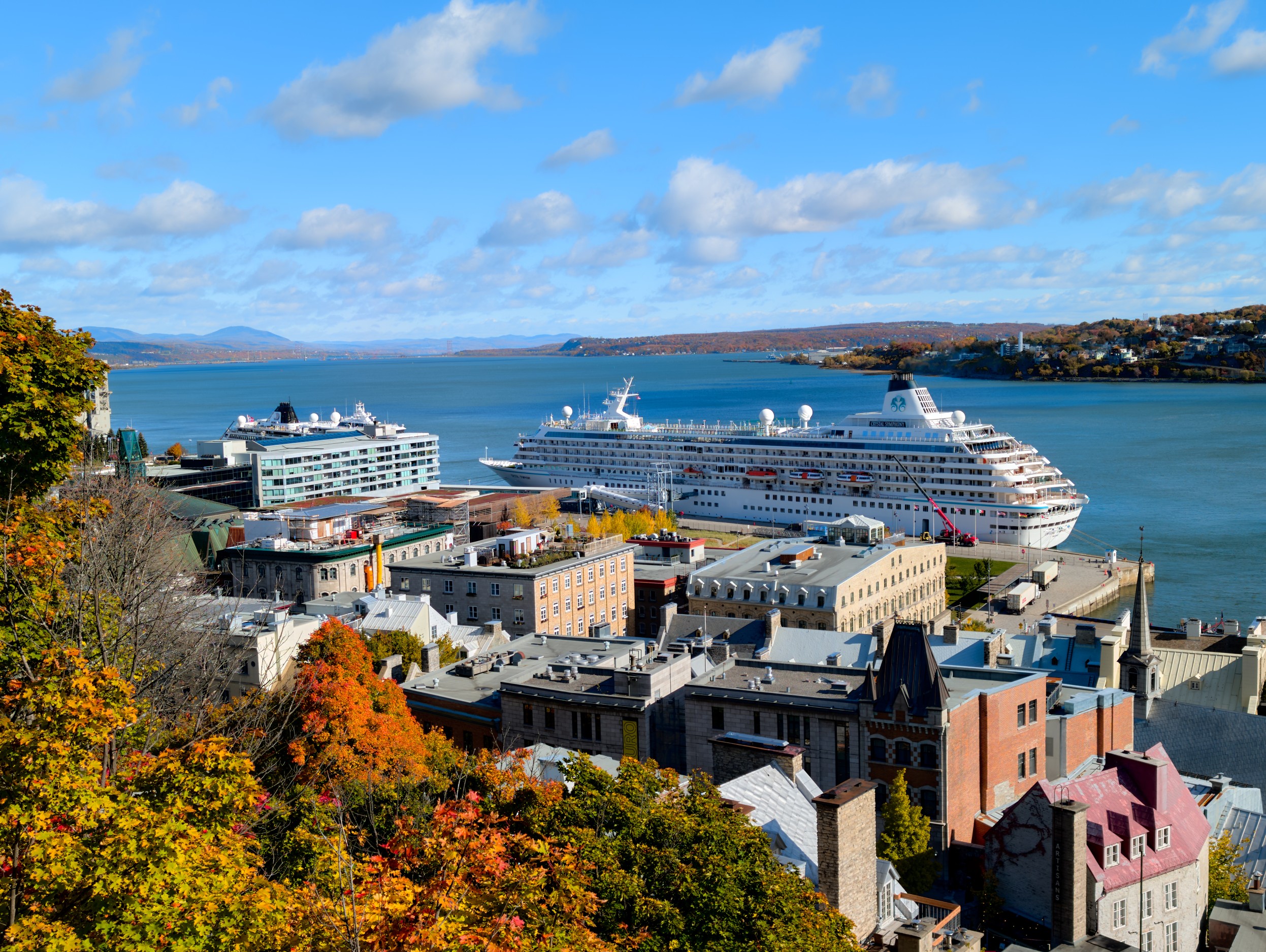 Vermont and Quebec 51 Elevated view across orange treetops and dense old-town roofs toward white passenger vessels moored along a wide river.